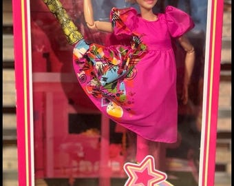 Puede incluir: Muñeca Barbie de "The Movie" con un vestido rosa y falda estampada, con peinado rubio. La muñeca está en una caja transparente con el logo de Barbie y un diseño en forma de estrella.