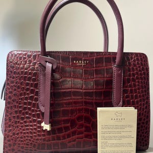 May include: A burgundy leather handbag with a crocodile skin texture. The bag has two handles and a small gold-colored dog charm. The brand name "RADLEY LONDON" is visible on the front. A tag with care instructions is attached.
