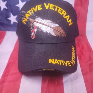 May include: A black baseball cap featuring yellow embroidered text that reads NATIVE VETERAN across the front and brim. An embroidered graphic of a white and brown feather with red and white accents is positioned above the brim on the cap.