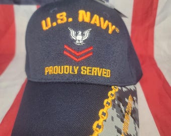 May include: A navy blue baseball cap with the text "U.S. NAVY" in gold lettering. The cap features an eagle emblem and the words "PROUDLY SERVED". The brim has a camouflage pattern with gold chain detailing.