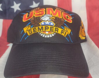 May include: Black baseball cap featuring the text "USMC" in red and gold, with an eagle emblem and the words "Semper Fi" in yellow. The cap has a curved brim and is set against an American flag background.