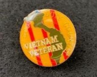 May include: A round enamel pin featuring a yellow background with three vertical red stripes. An olive green silhouette of the country of Vietnam is centered on the pin. The text VIETNAM VETERAN is printed in white across the center of the pin.
