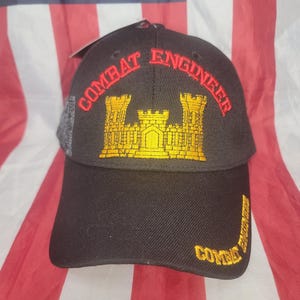 May include: Black baseball cap with "COMBAT ENGINEER" embroidered in red and gold. The cap features a gold castle emblem and additional text on the side. The background is an American flag.