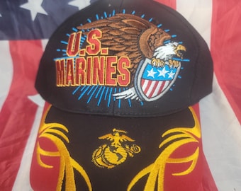 May include: Black baseball cap featuring an embroidered eagle with a shield and the text "U.S. MARINES" in red and blue. The cap's brim displays the Marine Corps emblem in gold. The background is an American flag.