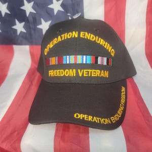 May include: Black baseball cap with the text "OPERATION ENDURING FREEDOM VETERAN" in gold lettering. The cap features a row of embroidered ribbons and is set against an American flag.