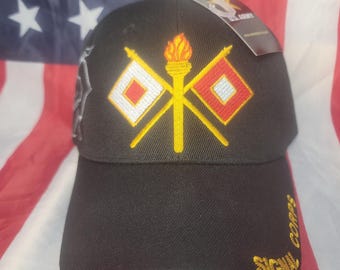 May include: Black baseball cap featuring a gold embroidered design of a torch and crossed flags. The words "SIGNAL CORPS" are embroidered in gold on the side. The cap is set against an American flag background.