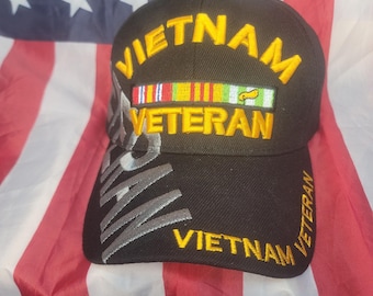 May include: Black baseball cap with "VIETNAM VETERAN" embroidered in yellow and silver. Features a colorful ribbon design. The cap is set against an American flag background.