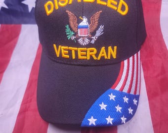 May include: A black baseball cap featuring the embroidered yellow text DISABLED VETERAN above an eagle emblem. The brim displays a partial American flag design with blue fabric, white stars, and red and white stripes on the right side.