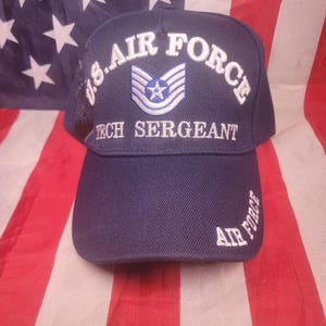 Pode incluir: Um boné de beisebol azul marinho com as palavras "U.S. AIR FORCE" e "TECH SERGEANT" bordadas em branco. O boné apresenta um emblema azul e branco. As palavras "AIR FORCE" também são bordadas na aba. O boné é exibido em frente a uma bandeira americana.
