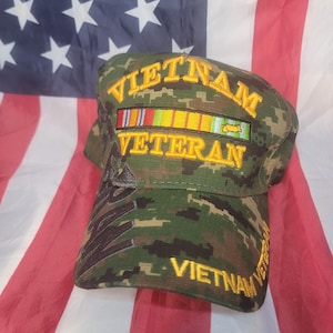 Könnte beinhalten: Eine Camouflage-Baseballkappe mit den goldfarbenen gestickten Worten "VIETNAM VETERAN". Die Kappe zeigt eine Reihe farbiger Bänder und ist vor einer amerikanischen Flagge platziert.
