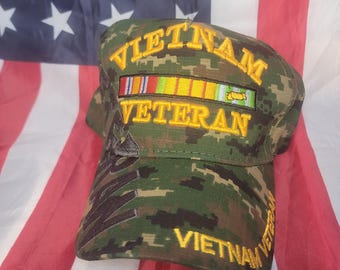 May include: A camouflage baseball cap with the words "VIETNAM VETERAN" embroidered in gold. The cap features a series of colored ribbons and is set against the backdrop of an American flag.