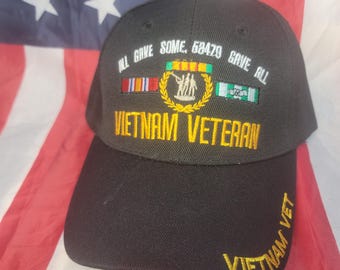 May include: Black baseball cap with embroidered text and patches. The cap reads "ALL GAVE SOME, 58479 GAVE ALL" and "VIETNAM VETERAN" in yellow. The cap also features military ribbons and a Vietnam War memorial emblem. The background is an American flag.