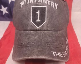 May include: Gray baseball cap with white embroidered text "1st INFANTRY" above a white outlined black shield with the number "1". The text "THE BIG RED ONE" is embroidered on the side. The cap is displayed on an American flag.