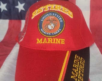 May include: A red baseball cap with the words "Retired" and "Marine" in gold lettering. The front of the cap features the United States Marine Corps emblem. The cap has a black brim with "Marine Retired" in gold.