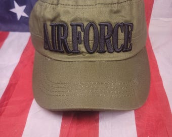 May include: An olive green military-style cadet cap featuring the word AIRFORCE embroidered in bold black capital letters across the front panel. The hat has a flat top, a structured crown, and a curved brim with visible stitching detail.
