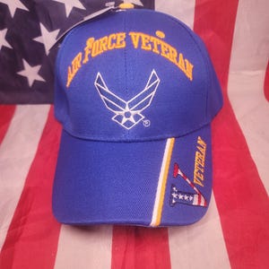 May include: A blue baseball cap with the words "Air Force Veteran" in gold lettering. The cap features the Air Force emblem and a design with the word "Veteran" on the side. The cap is displayed against an American flag.