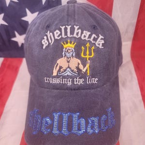 Peut inclure: Casquette de baseball bleu marine avec le mot "shellback" brodé en lettres gothiques blanches, une illustration d'un homme avec un trident et le texte "crossing the line". Le bord est brodé "shellback" en bleu. Accessoire décontracté.