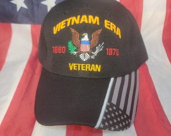 May include: Black baseball cap with "VIETNAM ERA" and "VETERAN" embroidered in gold. Features an eagle emblem and the years 1960 and 1975. The cap's side has a design of the American flag.