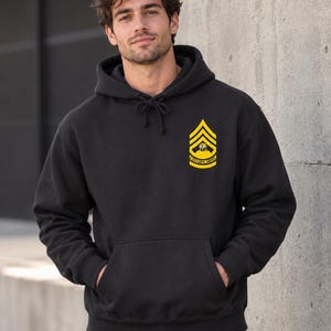 Puede incluir: Una sudadera con capucha negra que presenta un gráfico amarillo en el pecho izquierdo. El gráfico muestra una insignia de rango de galón con un símbolo de ojo en el centro y el texto "CYCLOPS ARMY" impreso en una cinta curva en la parte inferior.