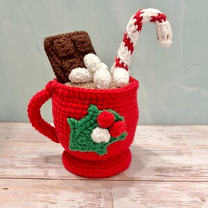 May include: A red crocheted mug filled with white marshmallows, a chocolate bar, and a red and white striped candy cane. The mug has a green holly and red berry design on the front. The mug is sitting on a wooden surface.