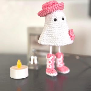 Könnte beinhalten: Eine handgefertigte weiße Häkel-Geisterfigur mit rosa Cowboyhut und Stiefeln. Der Geist hat schwarze Augen und eine rosa Blume. Eine kleine, weiße LED-Kerze mit gelber Flamme befindet sich im Vordergrund.