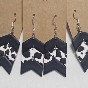 May include: Dangle earrings featuring a chevron-shaped design with three stacked segments. The top and bottom segments are solid matte black, while the middle segment features a black and white cow print pattern. The earrings hang from silver-toned hooks.