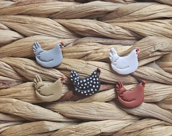 May include: A collection of six small, chicken-shaped ornaments in various colors, including gray, white, tan, black with white polka dots, and reddish-brown. Each chicken has a red comb and wattle. The ornaments are arranged on a woven, textured surface.