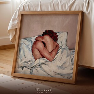 A square wood-framed print of the intimate figurative painting leaning on a wooden floor next to a bed. The warm sunlight creates a cozy atmosphere, showcasing the versatility of this modern art print for different room contexts like bedrooms or studios.