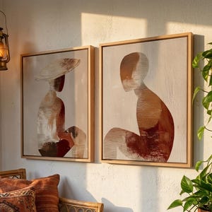 May include: Two framed abstract art pieces with neutral tones. The artwork features figures in shades of brown, beige, and red. The frames are light wood. The scene includes a rattan sofa with patterned pillows, a hanging lantern, and a potted plant.