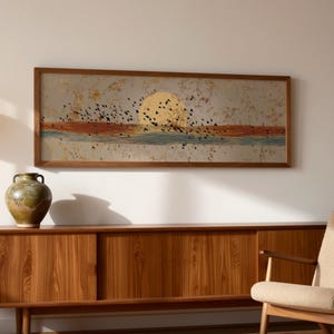 Puede incluir: Una impresión artística enmarcada representa un atardecer con un gran sol amarillo, pájaros y agua. La obra de arte cuelga sobre un mueble de madera con un jarrón y una silla moderna.