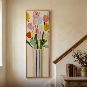 May include: A vertical rectangular framed painting featuring a bouquet of tulips in shades of pink, yellow, white, and blue, set in a striped vase. The artwork is displayed on a wall, with the text Trendart Studios printed at the bottom.