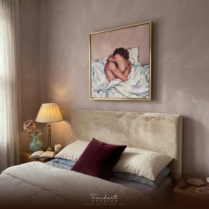 moody bedroom wall art above the bed in a dusty mauve room. A gold framed oil painting of a woman curled on white bedding is styled with a pleated lamp, a burgundy velvet pillow and fresh flowers.