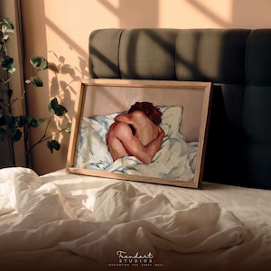 A horizontal framed print of a sensual modern art piece leaning against a dark gray headboard on a bed. Sunlight casts shadows on the wall, highlighting the warm beige and red tones of the impasto style figurative digital download for aesthetic home decor.