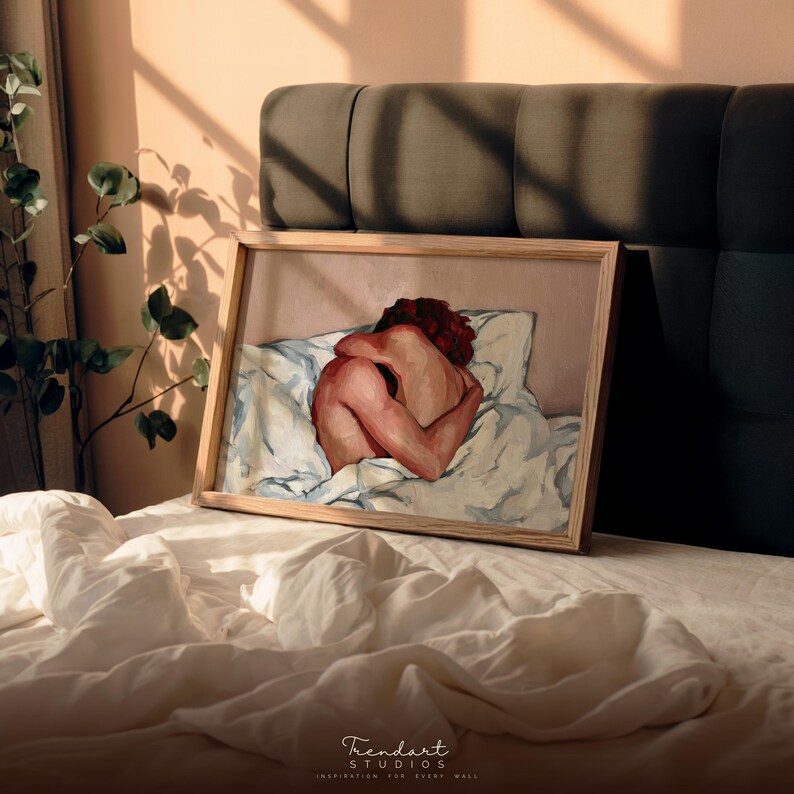 A horizontal framed print of a sensual modern art piece leaning against a dark gray headboard on a bed. Sunlight casts shadows on the wall, highlighting the warm beige and red tones of the impasto style figurative digital download for aesthetic home decor.