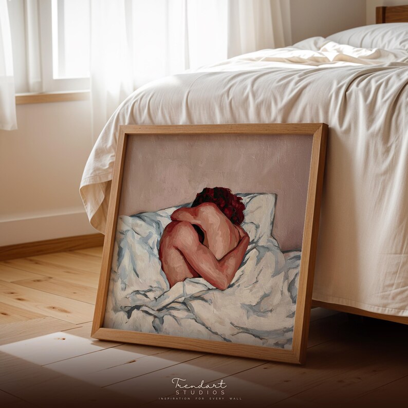 A square wood-framed print of the intimate figurative painting leaning on a wooden floor next to a bed. The warm sunlight creates a cozy atmosphere, showcasing the versatility of this modern art print for different room contexts like bedrooms or studios.