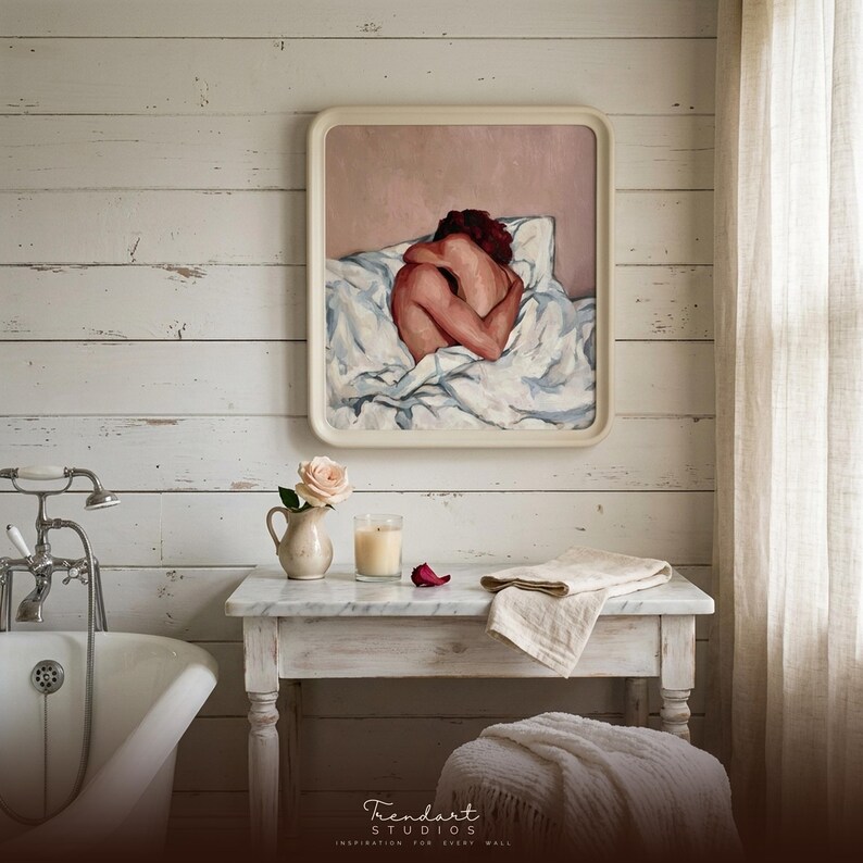coastal farmhouse bathroom wall art on whitewashed shiplap. A cream rounded frame holds a warm figure painting above a freestanding tub, a marble console, white roses and a flickering candle.