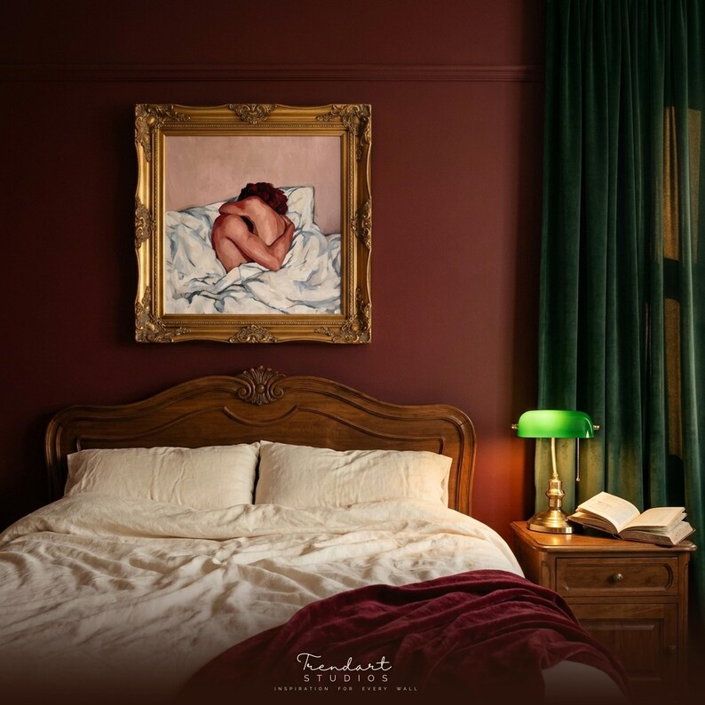 dark academia wall art for a moody bedroom. An ornate gold baroque frame holds a warm figurative oil painting on a deep maroon wall above a wooden headboard, a green banker lamp and an open book.