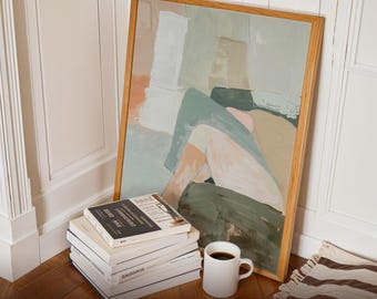 May include: A framed abstract art print with a light wood frame, leaning against a white wall. A stack of books and a white mug of coffee sit in front of the artwork. The art features muted tones of green, beige, and white.