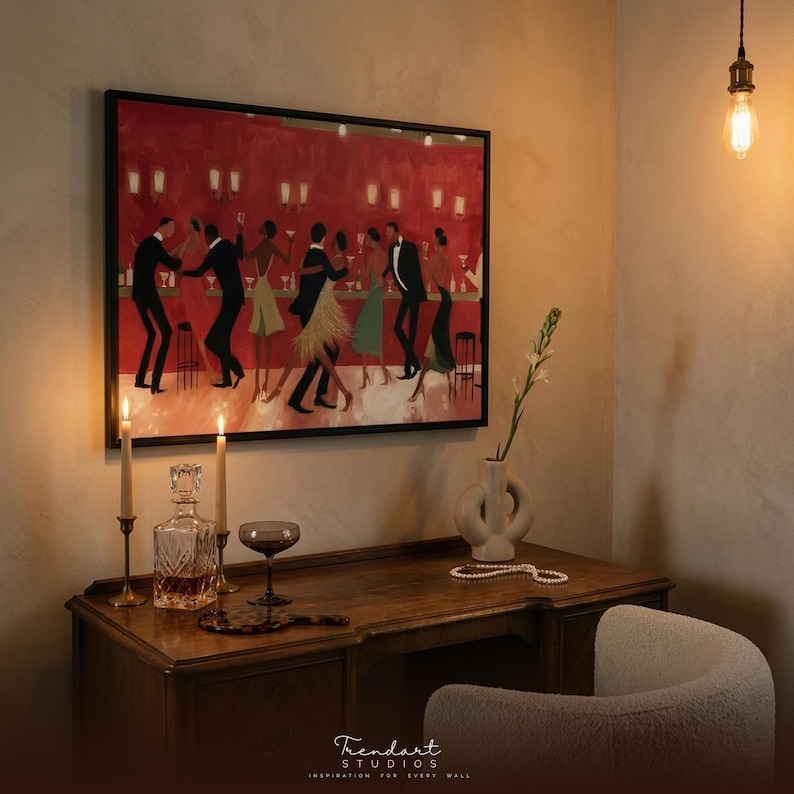 May include: A framed print shows a jazz club scene with dancing figures against a red backdrop. The artwork hangs above a wooden desk with a decanter, glass, candles, and a vase. A pearl necklace and a white chair are also visible.
