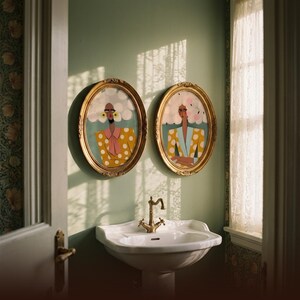 Funny bathroom wall art — two women prints in ornate gold oval frames above white pedestal sink, sage green powder room walls, natural window light with lace curtain