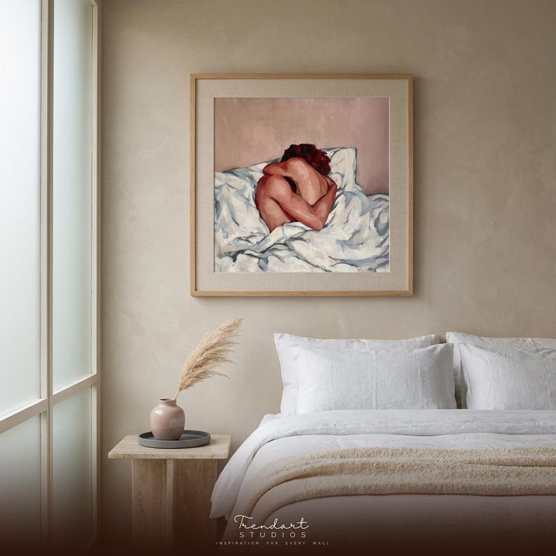 neutral bedroom wall art above a calm bed. A light wood frame with a white mat surrounds a soft figure oil painting, styled with a cream throw blanket and pampas grass in a round nightstand vase.