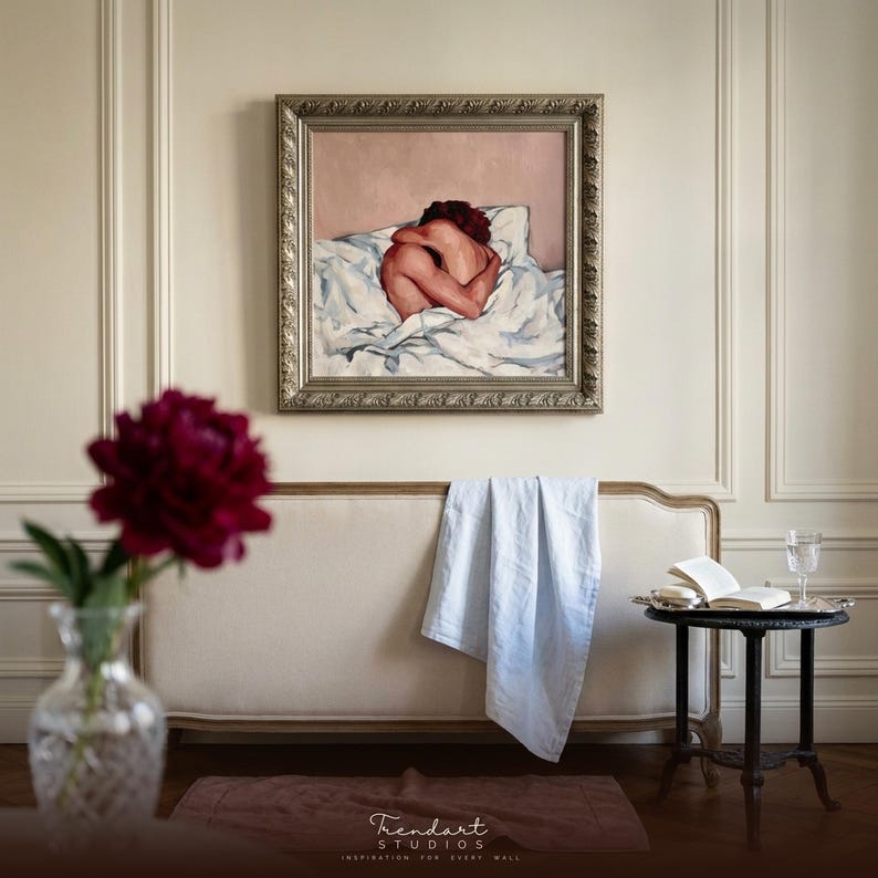 french country wall art on a cream paneled wall. A champagne ornate frame surrounds a sensual figure painting above a neutral bench, a deep red peony in a crystal vase and a small round side table.
