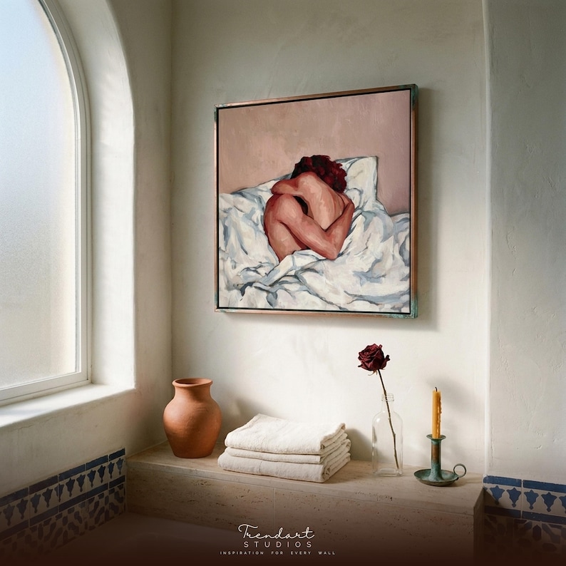 mediterranean bathroom wall art in a white arched alcove. A framed figurative painting in warm terracotta tones hangs above a niche with stacked towels, a clay vase, a single dried rose and blue patterned tiles.