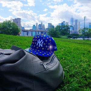 May include: A blue baseball cap with the New York Yankees logo, adorned with colorful circular designs, rests on a gray bag. The scene is set on a grassy hill with a city skyline and a bridge in the background.