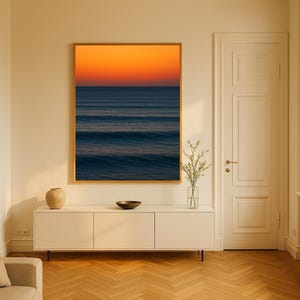 May include: A framed art print depicting an ocean scene with a vibrant orange and blue sunset. The artwork hangs above a white cabinet with a vase, bowl, and decorative items. The room features a wooden floor and a neutral color palette.