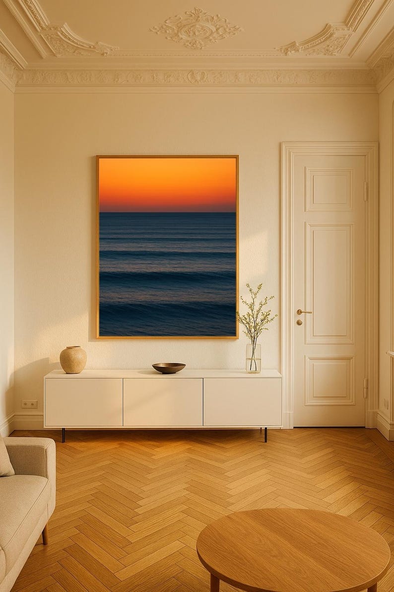 May include: A framed art print depicting an ocean scene with a vibrant orange and blue sunset. The artwork hangs above a white cabinet with a vase, bowl, and decorative items. The room features a wooden floor and a neutral color palette.