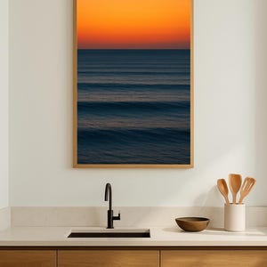 May include: A framed print of an ocean scene with a vibrant orange and blue gradient sky. The artwork hangs above a kitchen sink with wooden cabinets, a black faucet, and a white countertop. Wooden utensils are in a white ceramic holder.