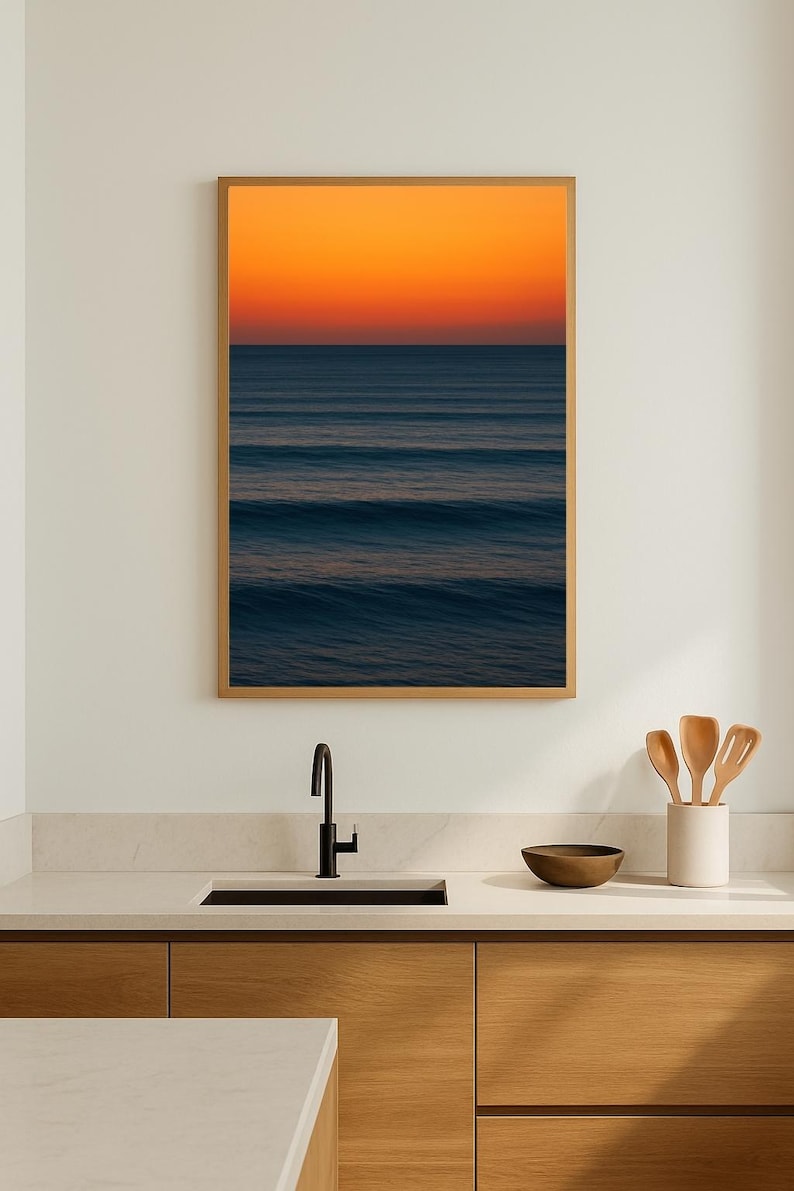 May include: A framed print of an ocean scene with a vibrant orange and blue gradient sky. The artwork hangs above a kitchen sink with wooden cabinets, a black faucet, and a white countertop. Wooden utensils are in a white ceramic holder.