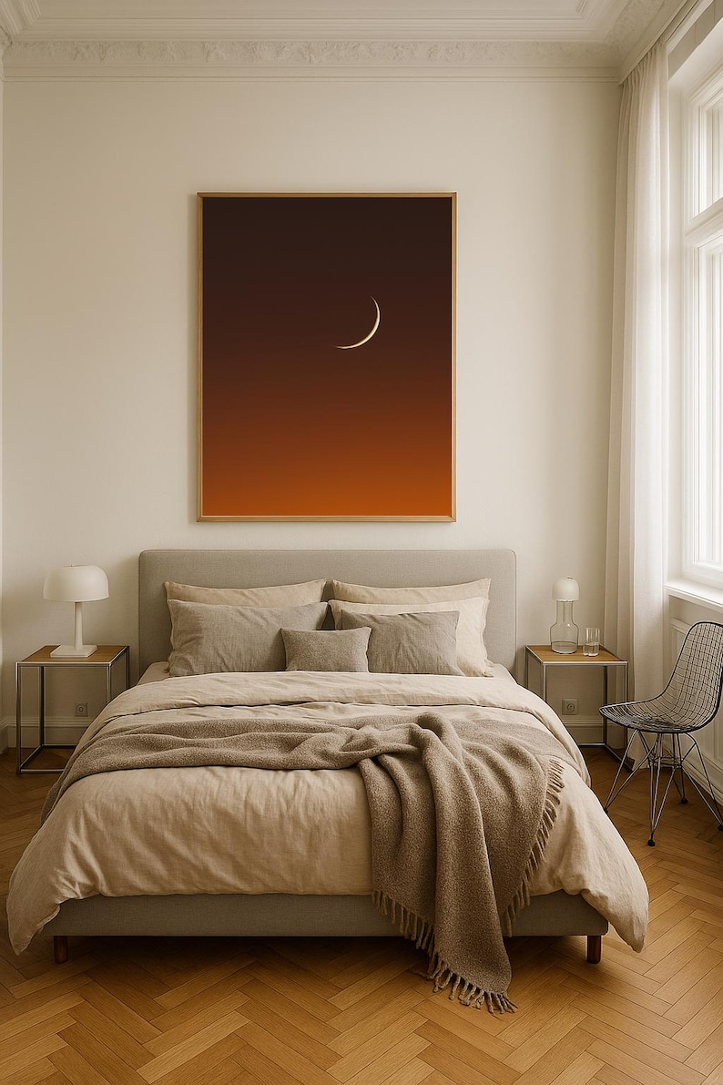 May include: A bedroom scene featuring a large framed print of a crescent moon against a gradient orange and brown sky. The bed is made with beige linens and a brown throw blanket. Two bedside tables with lamps flank the bed. A black wire chair sits near a window.