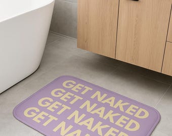 Puede incluir: Una alfombra de baño rectangular de color lavanda con las palabras "GET NAKED" repetidas cuatro veces en amarillo claro. La alfombra está colocada sobre un suelo embaldosado en un baño.